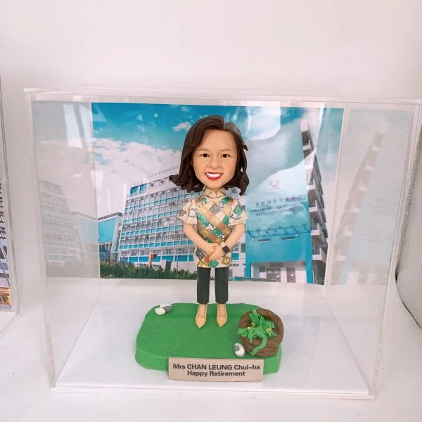 透明壓克力展示盒內，一尊女性人像公仔站在綠色草地質感底座上，短至中長髮、微笑，穿彩色幾何花紋上衣、深色及踝褲與米黃色鞋，雙手於身前交握。底座右側有一小簍綠植與白色小石裝飾。前方金色名牌刻有「Mrs CHAN LEUNG Chui-ha Happy Retirement」。背板是一張校園建築照片，左側為藍白相間的多層教學大樓，右側藍色牆面印有英文與中文校名「SHAU KEI WAN GOVERNMENT SECONDARY SCHOOL／筲箕灣官立中學」及學校盾形校徽，藍天白雲作背景。