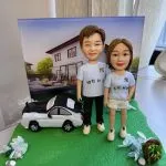 草綠不規則底座上的情侶人像公仔：左側男性穿淺藍T恤與黑長褲、白色運動鞋；右側女性穿同色系T恤搭白色短褲與白鞋。兩人並肩站立，身後擺放黑白配色的私家車模型；背景立起一幅現代獨棟住宅庭院的畫布，看似作為情境背板。底座兩側有藍白花材與小石頭、右前方放有裝綠葉的棕色小籃。