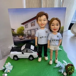 草綠不規則底座上的情侶人像公仔:左側男性穿淺藍T恤與黑長褲、白色運動鞋;右側女性穿同色系T恤搭白色短褲與白鞋。兩人並肩站立,身後擺放黑白配色的私家車模型;背景立起一幅現代獨棟住宅庭院的畫布,看似作為情境背板。底座兩側有藍白花材與小石頭、右前方放有裝綠葉的棕色小籃。