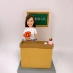教室場景的人像公仔：一位短髮女性公仔站在黃褐色講台後方，右手捧著橙紅色花束，左側講台上放著粉筆，右側是一個粉色筆筒插著彩色筆。背後掛著綠色小黑板，上寫白字「杏壇之光」。整體置於灰色階梯式底座，背景為素白牆。