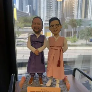 兩位男性人像公仔並肩站在同一灰色六角石磚紋底座上：左側公仔留短髮、淡鬍，穿紫色韓式長袍配棕色皮鞋，雙手於身前交握微笑；右側公仔戴眼鏡，穿粉色韓式長袍配白色運動鞋，右手叉腰、左手自然下垂微笑。底座前方放置橙色銘牌，英文字訊息以“Dear Steve”開頭，並署名“Danny (on behalf of APAC ClinOps team)”，內容表達感謝與祝福。