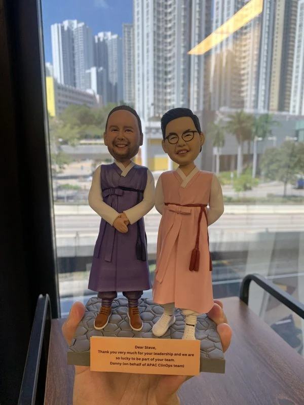 兩位男性人像公仔並肩站在同一灰色六角石磚紋底座上：左側公仔留短髮、淡鬍，穿紫色韓式長袍配棕色皮鞋，雙手於身前交握微笑；右側公仔戴眼鏡，穿粉色韓式長袍配白色運動鞋，右手叉腰、左手自然下垂微笑。底座前方放置橙色銘牌，英文字訊息以“Dear Steve”開頭，並署名“Danny (on behalf of APAC ClinOps team)”，內容表達感謝與祝福。