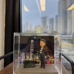 一款人像公仔置於透明展示盒中，背景為維多利亞港夜景與高樓天際線；地台為灰色石板路紋理。公仔穿深色西裝、白襯衫與條紋領帶，右手拿外帶咖啡杯，站姿輕鬆。腳邊擺放一台深綠色雙層電車模型；背景板可見英文字樣與標誌。整體放在木層板上，窗外是城市高樓景致。