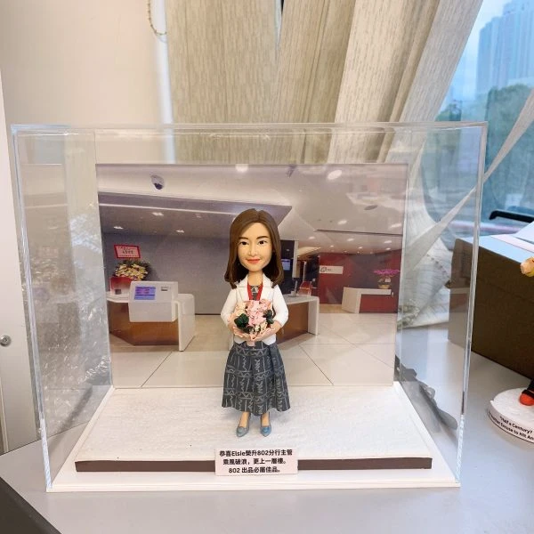 透明展示盒中的女性人像公仔：公仔留中長髮，穿白色外套、圖紋長裙與藍色鞋，胸前掛紅色頸繩識別證，雙手捧著粉色系花束微笑站立。背板為現代化室內大廳／櫃檯場景，地台為白色細沙質感，前方白色小牌印有「恭喜…分行主管」等祝賀字句。整體呈現升職慶賀的人像公仔擺設。