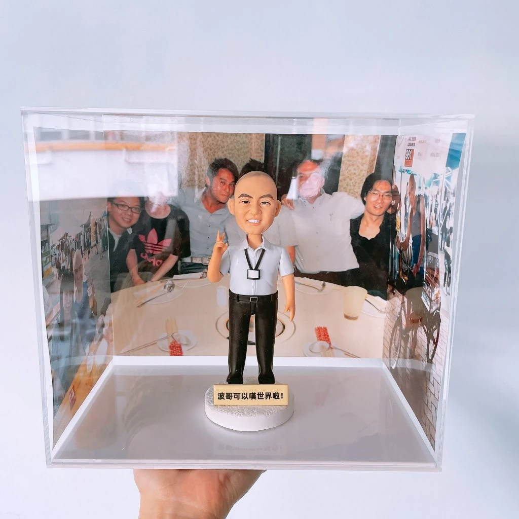 透明壓克力展示盒中的退休主題人像公仔：禿髮男性公仔穿淺灰短袖襯衫、黑長褲與黑皮鞋，胸前掛識別證，右手比出V字手勢微笑站立；圓形白色底座前方有金色小牌，刻有中文鼓勵字句。展示盒三面以聚餐與活動照片做背景，呈現同事情誼與回憶。整體乾淨明亮，適合作為退休公仔紀念擺設。