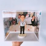 透明壓克力展示盒中的退休主題人像公仔：禿髮男性公仔穿淺灰短袖襯衫、黑長褲與黑皮鞋，胸前掛識別證，右手比出V字手勢微笑站立；圓形白色底座前方有金色小牌，刻有中文鼓勵字句。展示盒三面以聚餐與活動照片做背景，呈現同事情誼與回憶。整體乾淨明亮，適合作為退休公仔紀念擺設。