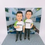 兩位男性人像公仔並排站立於灰色石磚紋底座，穿白色與綠色拼接的 Deloitte. 品牌T恤與黑色運動長褲、黑色運動鞋；左側公仔比出大拇指，右側公仔一手插袋。背板為室內中庭辦公空間照片，牆面與上方招牌可見「Deloitte. 德勤」字樣，整體呈現企業品牌與團隊氛圍的人像公仔場景。