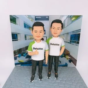 兩位男性人像公仔並排站立於灰色石磚紋底座,穿白色與綠色拼接的 Deloitte. 品牌T恤與黑色運動長褲、黑色運動鞋;左側公仔比出大拇指,右側公仔一手插袋。背板為室內中庭辦公空間照片,牆面與上方招牌可見「Deloitte. 德勤」字樣,整體呈現企業品牌與團隊氛圍的人像公仔場景。