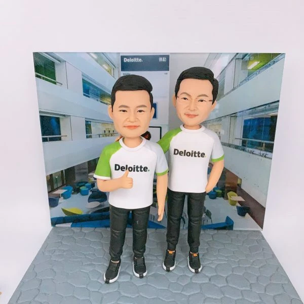 兩位男性人像公仔並排站立於灰色石磚紋底座，穿白色與綠色拼接的 Deloitte. 品牌T恤與黑色運動長褲、黑色運動鞋；左側公仔比出大拇指，右側公仔一手插袋。背板為室內中庭辦公空間照片，牆面與上方招牌可見「Deloitte. 德勤」字樣，整體呈現企業品牌與團隊氛圍的人像公仔場景。