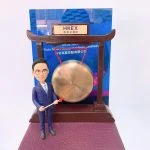 以香港交易所敲鑼儀式為主題的人像公仔：身穿深藍西裝與白襯衫的男性公仔，手持紅頭木槌站在銅鑼旁；銅鑼懸掛於深木色拱形架上，頂部銘牌印有「HKEX 香港交易所」。背板為藍色活動海報樣式，底座為暗紫色質感平台。整體呈現企業代表在港交所參與上市敲鑼的慶賀場景，作為金融領域的紀念人像公仔。