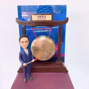 以香港交易所敲鑼儀式為主題的人像公仔：身穿深藍西裝與白襯衫的男性公仔，手持紅頭木槌站在銅鑼旁；銅鑼懸掛於深木色拱形架上，頂部銘牌印有「HKEX 香港交易所」。背板為藍色活動海報樣式，底座為暗紫色質感平台。整體呈現企業代表在港交所參與上市敲鑼的慶賀場景，作為金融領域的紀念人像公仔。