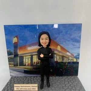 退休致敬主題的人像公仔:黑色長髮女性,身穿全黑制服外套與長褲、黑色長靴,雙手交叉自信站姿,面帶微笑。腳下為灰色石板紋地台,前方有英文字牌:「Happy Retirement, Melon! We will miss you! ABU」。背板為夜色中的 McDonald’s 麥當勞門店外觀,招牌金色拱門清晰可見。整體呈現餐飲門店管理人員的榮休祝福禮物場景。