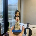 同事退休主題的人像公仔：女同事長髮微笑，穿白色外套與多層荷葉邊長裙，手持紅色禮盒站立；深藍色斜角名牌座刻有中文祝福文字。背景為辦公室窗景與城市大樓，整體呈現精緻小巧、可單手托起的桌上型退休禮物的人像公仔。