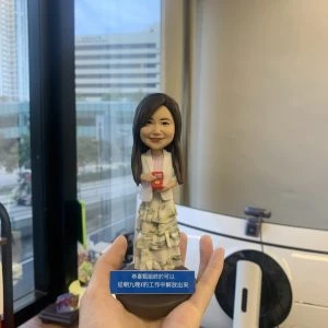 同事退休主題的人像公仔：女同事長髮微笑，穿白色外套與多層荷葉邊長裙，手持紅色禮盒站立；深藍色斜角名牌座刻有中文祝福文字。背景為辦公室窗景與城市大樓，整體呈現精緻小巧、可單手托起的桌上型退休禮物的人像公仔。