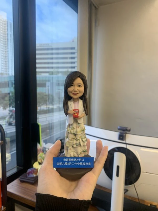 同事退休主題的人像公仔：女同事長髮微笑，穿白色外套與多層荷葉邊長裙，手持紅色禮盒站立；深藍色斜角名牌座刻有中文祝福文字。背景為辦公室窗景與城市大樓，整體呈現精緻小巧、可單手托起的桌上型退休禮物的人像公仔。