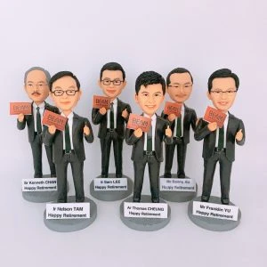 六位男性人像公仔並排站立的退休主題擺設：皆著黑色西裝、白襯衫與領帶，右手比讚，左手拿著橘紅色「BEAM」字樣卡片；每個公仔腳前的圓形底座附白色名牌，分別印有「Sr Kenneth CHAN」「Ir Nelson TAM」「Ir Sam LEE」「Ar Thomas CHEUNG」「Mr Benny AU」「Mr Franklin YU」及「Happy Retirement」。背景為純白，整體呈現團隊向資深成員致敬的退休人像公仔組合。