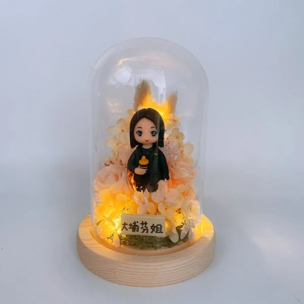 一個人像公仔被置於透明玻璃圓頂的永生花花罩內，底座為淺色木座。公仔留中長直髮，穿深色外套，手持橙色奶瓶造型道具；周圍以粉橘與米色系的永生花、繡球與乾燥芒草環繞，內置暖黃小燈點亮整體。前方有小牌寫著「大埔芳姐」。