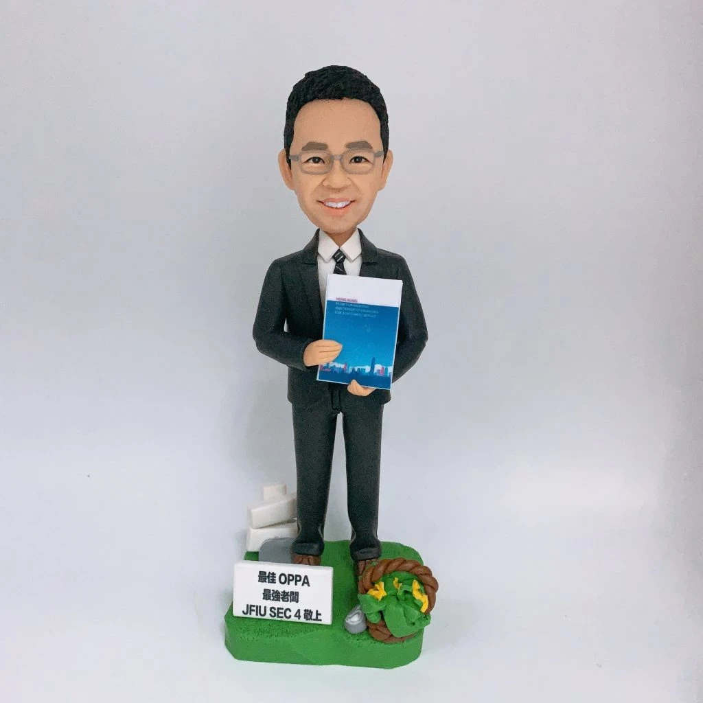 穿深灰色西裝、打條紋領帶的男性人像公仔，戴眼鏡、微笑站立，雙手於身前捧著一本藍色封面的文件冊。公仔置於草綠色地台，腳邊有白色指示牌造型與小型告示牌（印有「最佳 OPPA 最高老闆 JFIU SEC 4 以上」字樣），右側點綴麻繩圈與黃色小花及銀色小物件。背景為純白色，整體呈現尊敬老闆與退休致意的禮贈氛圍。
