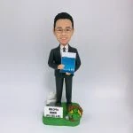 穿深灰色西裝、打條紋領帶的男性人像公仔，戴眼鏡、微笑站立，雙手於身前捧著一本藍色封面的文件冊。公仔置於草綠色地台，腳邊有白色指示牌造型與小型告示牌（印有「最佳 OPPA 最高老闆 JFIU SEC 4 以上」字樣），右側點綴麻繩圈與黃色小花及銀色小物件。背景為純白色，整體呈現尊敬老闆與退休致意的禮贈氛圍。