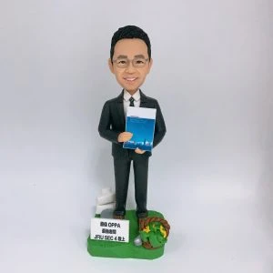 穿深灰色西裝、打條紋領帶的男性人像公仔，戴眼鏡、微笑站立，雙手於身前捧著一本藍色封面的文件冊。公仔置於草綠色地台，腳邊有白色指示牌造型與小型告示牌（印有「最佳 OPPA 最高老闆 JFIU SEC 4 以上」字樣），右側點綴麻繩圈與黃色小花及銀色小物件。背景為純白色，整體呈現尊敬老闆與退休致意的禮贈氛圍。