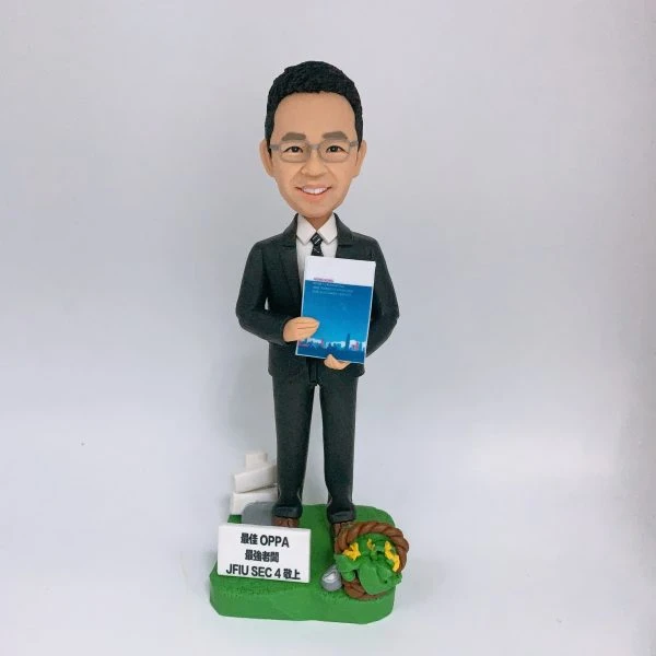 穿深灰色西裝、打條紋領帶的男性人像公仔，戴眼鏡、微笑站立，雙手於身前捧著一本藍色封面的文件冊。公仔置於草綠色地台，腳邊有白色指示牌造型與小型告示牌（印有「最佳 OPPA 最高老闆 JFIU SEC 4 以上」字樣），右側點綴麻繩圈與黃色小花及銀色小物件。背景為純白色，整體呈現尊敬老闆與退休致意的禮贈氛圍。