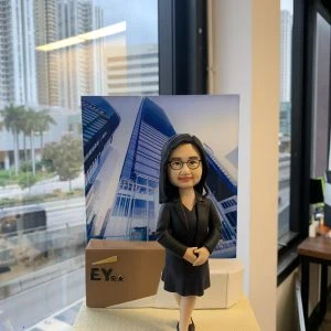 人像公仔以專業女性形象呈現:黑色套裝與裙裝、配戴眼鏡與項鍊,雙手交疊站姿,自信微笑。背板為現代玻璃寫字樓外觀圖片;左側有帶有 EY 標誌與中文字「安永」的立方體裝飾。底座前方小牌以英文寫上對退休的感謝與祝福語。場景位於室內窗邊,可見城市高樓與街景。