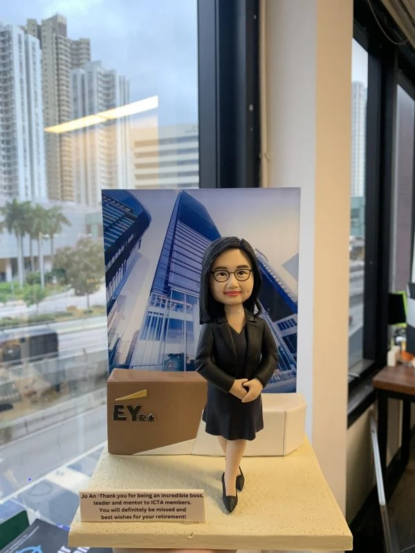 人像公仔以專業女性形象呈現：黑色套裝與裙裝、配戴眼鏡與項鍊，雙手交疊站姿，自信微笑。背板為現代玻璃寫字樓外觀圖片；左側有帶有 EY 標誌與中文字「安永」的立方體裝飾。底座前方小牌以英文寫上對退休的感謝與祝福語。場景位於室內窗邊，可見城市高樓與街景。