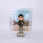 一款人像公仔置於透明展示盒內，背景是色彩繽紛的主題樂園布景（玩具造型與字母積木裝飾）。公仔戴黑框眼鏡，穿深色連帽外套與卡其長褲、黑白運動鞋，胸前配有圓形名牌，雙臂交叉站立在灰色底座上。底座金屬牌刻有「Toystoryland • Grizzly • Adventureland Manager Antony」等字樣。