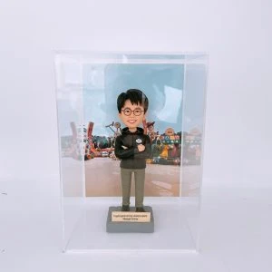 一款人像公仔置於透明展示盒內,背景是色彩繽紛的主題樂園布景(玩具造型與字母積木裝飾)。公仔戴黑框眼鏡,穿深色連帽外套與卡其長褲、黑白運動鞋,胸前配有圓形名牌,雙臂交叉站立在灰色底座上。底座金屬牌刻有「Toystoryland • Grizzly • Adventureland Manager Antony」等字樣。