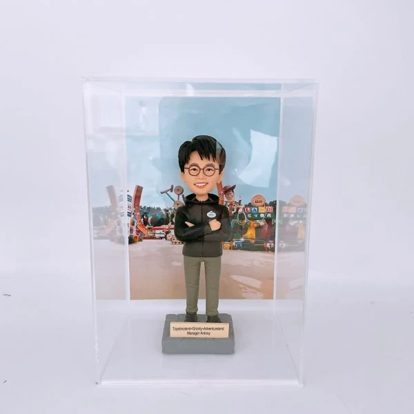 一款人像公仔置於透明展示盒內，背景是色彩繽紛的主題樂園布景（玩具造型與字母積木裝飾）。公仔戴黑框眼鏡，穿深色連帽外套與卡其長褲、黑白運動鞋，胸前配有圓形名牌，雙臂交叉站立在灰色底座上。底座金屬牌刻有「Toystoryland • Grizzly • Adventureland Manager Antony」等字樣。