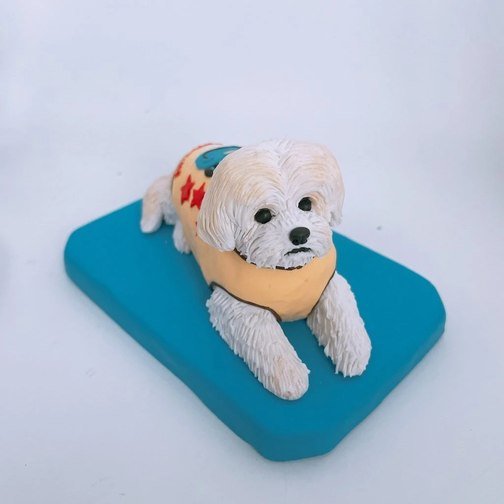 白色馬爾濟斯造型的寵物公仔以趴臥姿勢置於藍色長方形底座上，毛髮細緻呈現柔軟捲蓬質感；圓大黑眼與黑色鼻頭突出。公仔身穿淺黃色背心，背部裝飾紅色星星與藍色圓形圖案，前雙掌向前伸直、後腿收於身側，整體給人溫馴可愛的觀感。