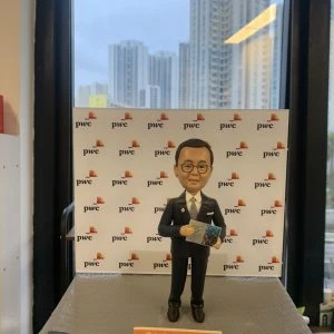 人像公仔站在灰色台座上,身穿深色西裝與領帶,配戴眼鏡,手持一本印有城市景觀圖的冊子;背板重複出現「pwc」與彩色方塊標誌,前緣橘色小牌寫有祝福語與年份2024;場景位於室內窗前,可見遠處高樓天際線。整體呈現專業商務與顧問氛圍的人像公仔展示。