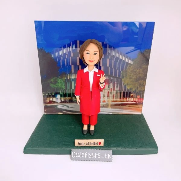 城市背景的送別主題人像公仔：短棕髮女性公仔身穿亮紅色套裝與白襯衫，右手比V手勢，腳踩黑色皮鞋，站在深綠色紋理底座上。身後為彩色城市建築背板，前方小木牌寫有「Eunice, All the Best ❤️」，底座前緣放置「Cutefigure_hk」標籤。背景為純白。