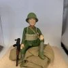 一款軍事風格的人像公仔坐在米色沙包堆上，身穿綠色軍裝與同色頭盔，胸前配有彈藥攜行具與斜揹背帶，領口見紅色領章細節；左側倚著黑棕色步槍道具。公仔面帶微笑，右膝抬起、左腿下垂，背景為淺色展示空間。