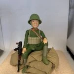 一款軍事風格的人像公仔坐在米色沙包堆上，身穿綠色軍裝與同色頭盔，胸前配有彈藥攜行具與斜揹背帶，領口見紅色領章細節；左側倚著黑棕色步槍道具。公仔面帶微笑，右膝抬起、左腿下垂，背景為淺色展示空間。