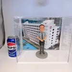 透明壓克力展示盒內，一尊男性人像公仔站在灰色圓形底座上，短髮、戴圓框眼鏡，穿淺灰短袖襯衫、黑色長褲與黑鞋，打深色點紋領帶，雙手於身前交握。背板是一幢白色長條形校舍大樓的照片，外牆有中文校名「慈南中學」及紅色圓形校徽，樓旁可見藍色戶外球場與籃球場標線。展示盒左側外面擺著一罐藍紅銀配色的百事可樂鋁罐作比例參考。