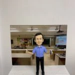 教室主題的人像公仔：短髮男性公仔身穿藍色短袖襯衫與黑色長褲，雙臂微張站立，右手持白色粉筆、左手拿書本。公仔置於顆粒感白色台座前，背板為教室場景，包含黑板、投影幕、講桌、實驗桌與牆上海報。