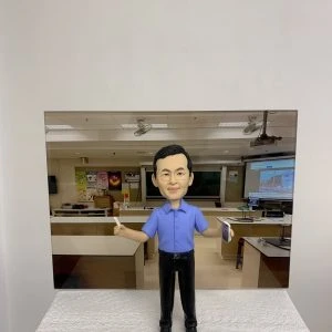 教室主題的人像公仔:短髮男性公仔身穿藍色短袖襯衫與黑色長褲,雙臂微張站立,右手持白色粉筆、左手拿書本。公仔置於顆粒感白色台座前,背板為教室場景,包含黑板、投影幕、講桌、實驗桌與牆上海報。