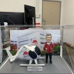 退休祝福主題的人像公仔場景：一位短灰髮、微笑的男性身穿紅色連帽外套、黑長褲與黑色運動鞋，雙手插袋站立；旁邊是一架白色直升機模型，機身與艙門上印有紅色圓形圖案與「LEE KUM KEE」字樣。兩者置於灰色地台，前方有英文銘牌：「Mr Terrence Lo — Happy Retirement and all the best to the new chapter」。背景背板為公司工廠大樓外觀與品牌標誌。整組作品收納在透明展示盒內，拍攝環境為室內辦公區與EXIT指示牌可見。整體呈現企業退休致意的人像公仔禮物。