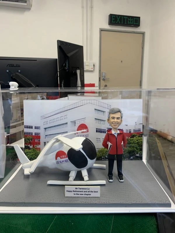 退休祝福主題的人像公仔場景：一位短灰髮、微笑的男性身穿紅色連帽外套、黑長褲與黑色運動鞋，雙手插袋站立；旁邊是一架白色直升機模型，機身與艙門上印有紅色圓形圖案與「LEE KUM KEE」字樣。兩者置於灰色地台，前方有英文銘牌：「Mr Terrence Lo — Happy Retirement and all the best to the new chapter」。背景背板為公司工廠大樓外觀與品牌標誌。整組作品收納在透明展示盒內，拍攝環境為室內辦公區與EXIT指示牌可見。整體呈現企業退休致意的人像公仔禮物。
