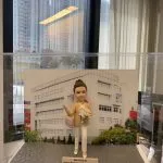 退休祝福主題的人像公仔：女性公仔盤髮造型，穿淺卡其外套、白襯衫與白色長褲，腳穿白鞋，右手揮手致意，左手抱粉白色花束，肩背菱格紋手袋。站於灰色石板紋底座，前方金色名牌刻有退休祝福語。背板為白色教學／辦公大樓與綠樹景觀，整體置於透明展示盒內，呈現優雅告別職涯的新章節。