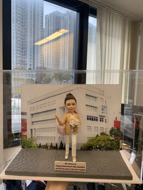 退休祝福主題的人像公仔：女性公仔盤髮造型，穿淺卡其外套、白襯衫與白色長褲，腳穿白鞋，右手揮手致意，左手抱粉白色花束，肩背菱格紋手袋。站於灰色石板紋底座，前方金色名牌刻有退休祝福語。背板為白色教學／辦公大樓與綠樹景觀，整體置於透明展示盒內，呈現優雅告別職涯的新章節。
