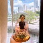 玻璃罩內的退休禮物人像公仔擺設：短捲髮、戴圓框眼鏡的女性公仔，身穿深色連衣裙與外罩格紋上衣，頸掛項鍊，站立於木質圓形底座上。玻璃罩內佈滿粉、白、紫色永生花與苔蘚，周圍點綴暖黃小燈串。前方小木牌寫有「Dear Ms. Lo, Happy Retirement!」。整體被手掌托起，背景為窗外城市高樓與綠樹景致。