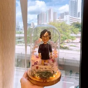 玻璃罩內的退休禮物人像公仔擺設:短捲髮、戴圓框眼鏡的女性公仔,身穿深色連衣裙與外罩格紋上衣,頸掛項鍊,站立於木質圓形底座上。玻璃罩內佈滿粉、白、紫色永生花與苔蘚,周圍點綴暖黃小燈串。前方小木牌寫有「Dear Ms. Lo, Happy Retirement!」。整體被手掌托起,背景為窗外城市高樓與綠樹景致。