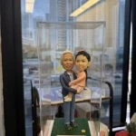 透明展示盒內的一組人像公仔：一位長者造型穿藍色外套、襯衫與長褲，雙手抱著一位穿粉色洋裝與髮飾的小朋友／bb公仔；兩人站在草地質感的綠色方形底座上，底座前有白色名牌印有祝福字樣。背景為室內窗邊，可見高樓與室內陳設的模糊景象。