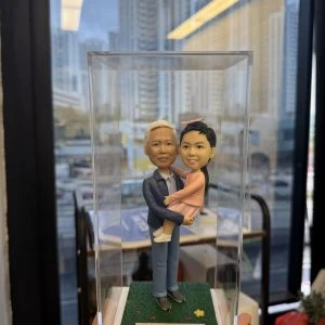 透明展示盒內的一組人像公仔：一位長者造型穿藍色外套、襯衫與長褲，雙手抱著一位穿粉色洋裝與髮飾的小朋友／bb公仔；兩人站在草地質感的綠色方形底座上，底座前有白色名牌印有祝福字樣。背景為室內窗邊，可見高樓與室內陳設的模糊景象。