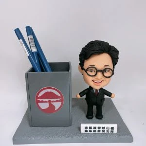 桌面擺設的人像公仔與筆筒組:短髮、戴黑色圓框眼鏡的男性Q版公仔,穿黑色西裝配深酒紅領帶,微笑站在灰色顆粒質感底座上。左側是一個灰色方形筆筒,正面貼有紅白圓形徽記圖案;筆筒內插著數支藍色筆。公仔前方放置白色小牌,上有多行黑色中文祝福字樣。背景為純白。