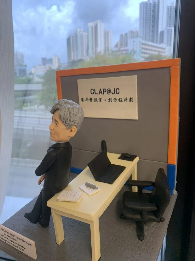 退休致敬主題的人像公仔：銀灰短髮男性穿黑色西裝與條紋領帶，側身倚靠米色長桌；桌上有開啟的筆電、筆與文件本、紙張疊。背板印有「CLAP@JC」與中文標語，台座前方附有英文名牌，標註社會服務相關職稱與退休祝福。整體置於室內窗邊場景中，可見城市高樓與藍天背景，呈現社福教育機構辦公氛圍的人像公仔。