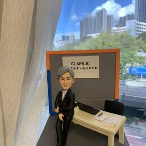 退休致敬主題的人像公仔：銀灰短髮男性穿黑色西裝與條紋領帶，側身倚靠米色長桌；桌上有開啟的筆電、筆與文件本、紙張疊。背板印有「CLAP@JC」與中文標語，台座前方附有英文名牌，標註社會服務相關職稱與退休祝福。整體置於室內窗邊場景中，可見城市高樓與藍天背景，呈現社福教育機構辦公氛圍的人像公仔。