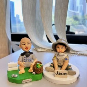桌面上的兩款小朋友主題人像公仔：左側bb坐在草綠底座，穿淺灰連身衣與深灰圍兜，嘴巴張開微笑，旁有白色木牌造型與小石頭、棕色編織籃內裝綠色葉菜；底座前緣貼有「CUTEFIGURE.HK」小牌。右側bb坐在白色圓底座，戴寬邊卡其漁夫帽、穿米色短袖與短褲，腳著綠色涼鞋，胸前圍巾寫「HANDSOME」，雙腳前放名牌「Jace」。背景為室內窗邊與柔紗簾、遠處高樓景色散景。